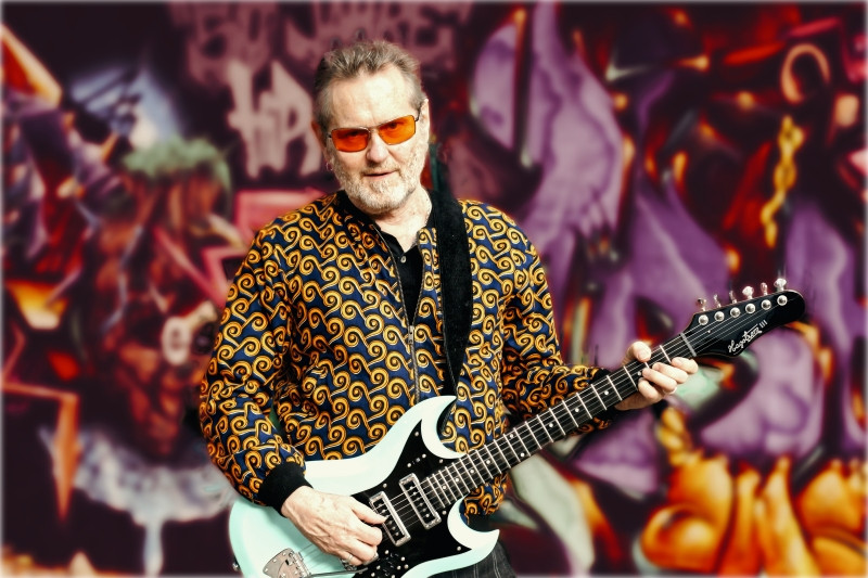 STOPPOK steht mit seiner weißen Gitarre vor einem lila-roten Hintergrund im Graffti-Stil, den Blick zur Kamera gerichtet.