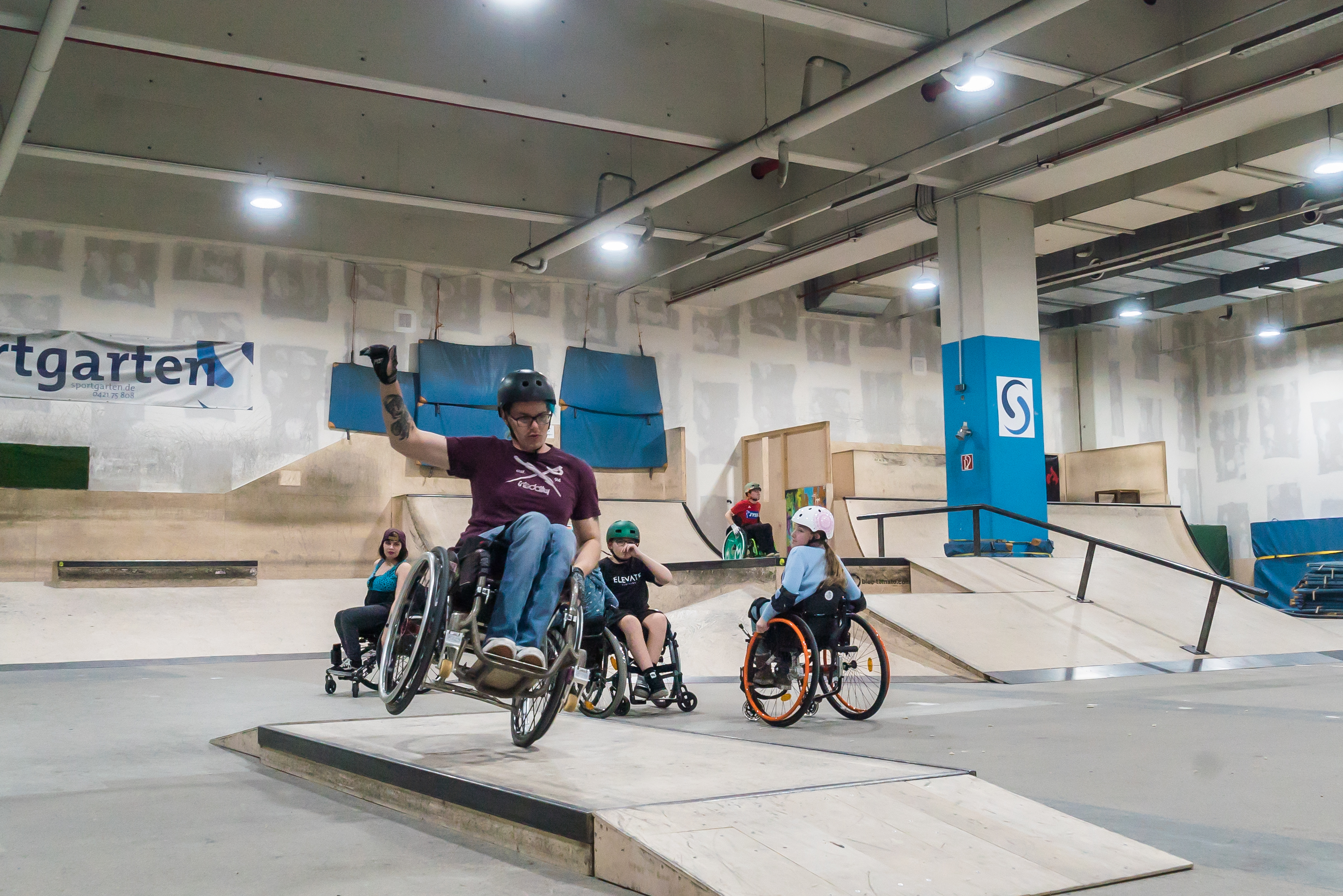 Fünf Rollstuhlfahrer:innen fahren in der Skatehalle am Sportgarten über Rampen und Pipes.