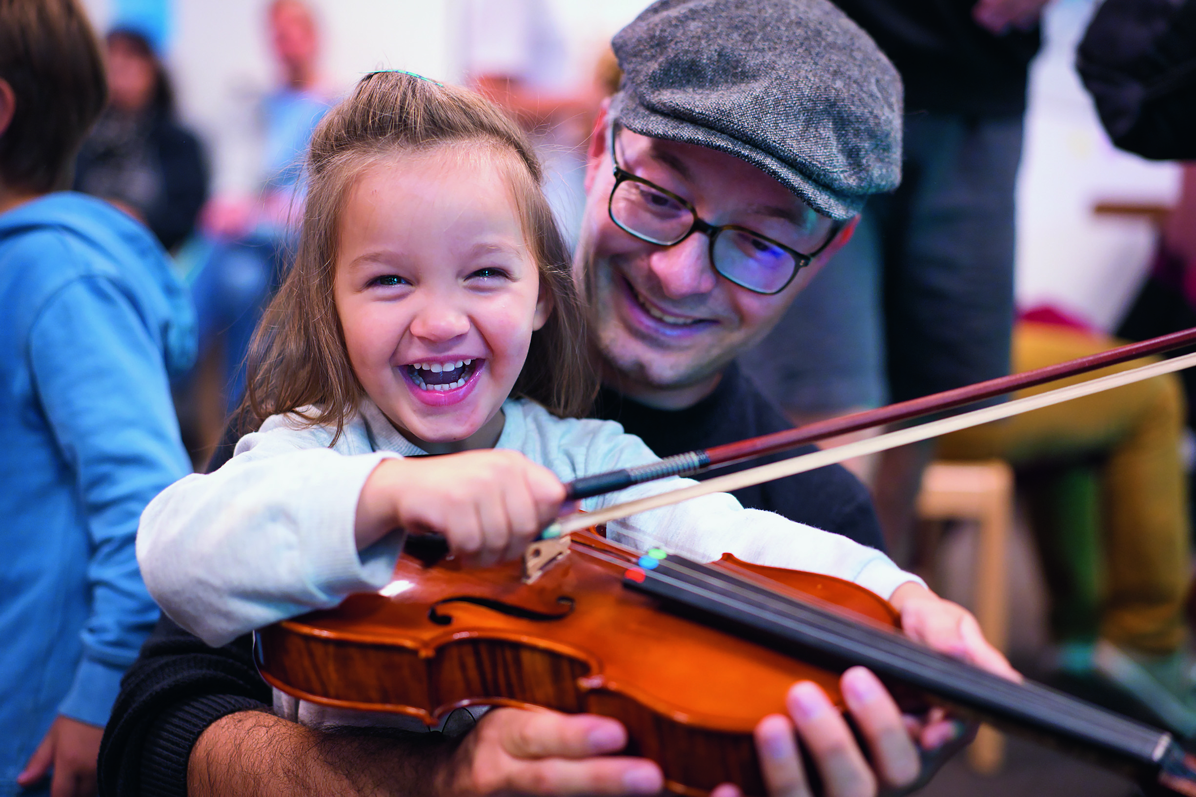 Ein Mädchen hält lachend den Bogen einer Bratsche, während ein Musiker der Bremer Philharmoniker ihr die Bratsche hält.
